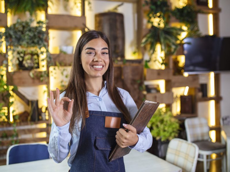 Happy beautiful smiling waitress wearing apron holding a folder menu in a restaurant, looking at camera, standing in cozy coffeehouse, good service