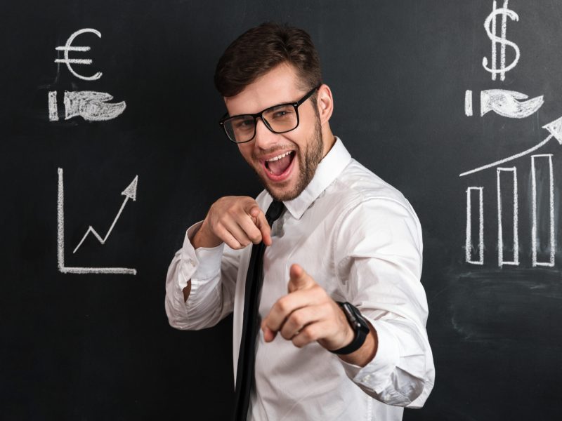 Young smiling handsome man in glasses pointing with two fingers on you, looking at camera, isolated over blackboard with financial statistics